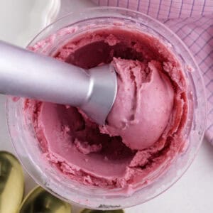 Une cuillère à glace en métal dans un récipient de crème glacée rose vif, avec un tissu à carreaux roses et des cuillères en or à proximité.