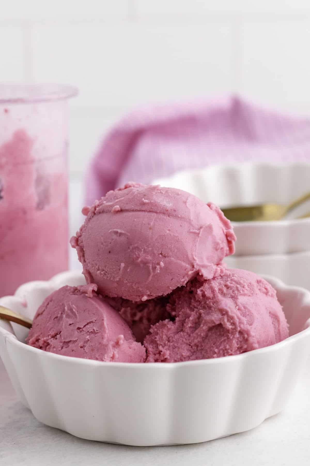 Trois boules de yogourt glacée aux fraises dans un bol blanc festonné, avec une cuillère dorée et une serviette rose en arrière-plan.