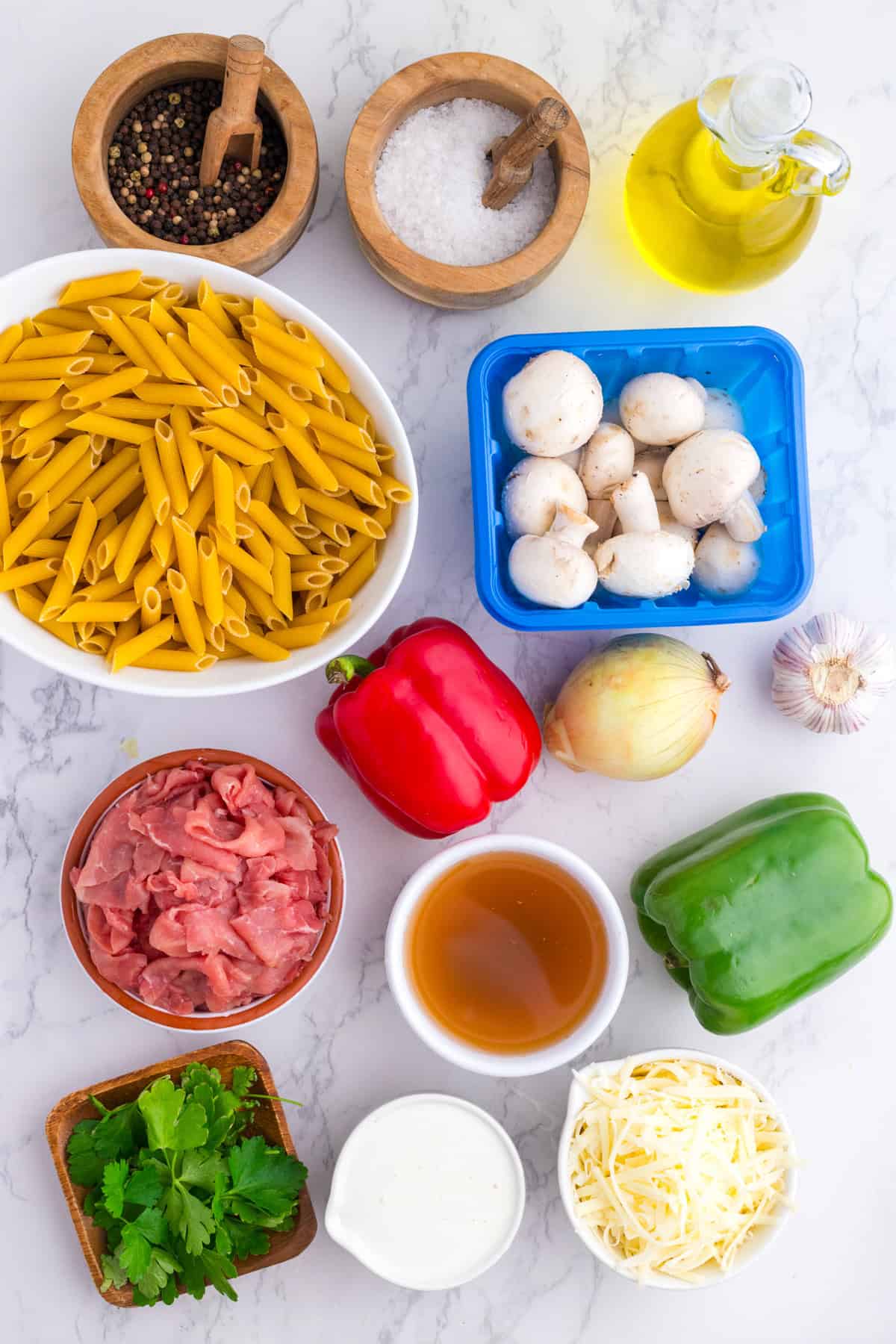 Top-down view of ingredients on a marble surface, including penne pasta, bell peppers, mushrooms, onion, garlic, raw meat, parsley, broth, cream, shredded cheese, seasonings, and olive oil.