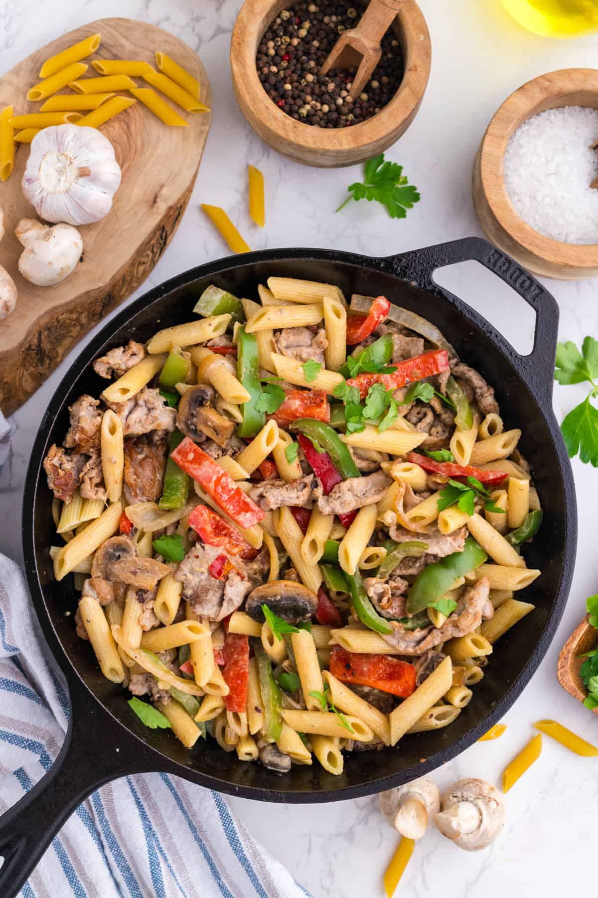 A cast iron skillet filled with penne pasta, sliced sausage, bell peppers, mushrooms, and fresh parsley sits on a marble countertop surrounded by cooking ingredients.