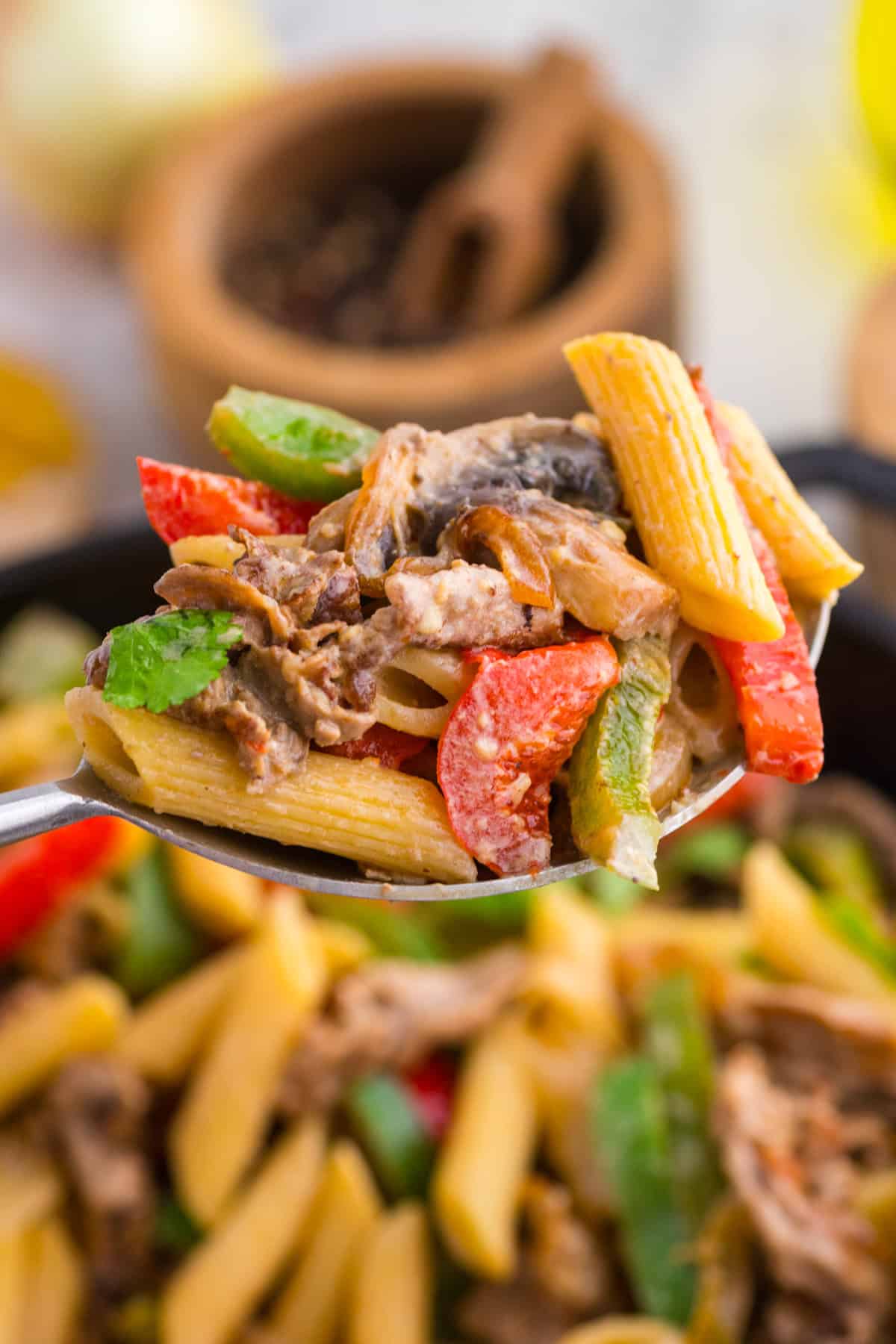 A close-up of a spoonful of penne pasta with sliced red and green bell peppers, mushrooms, and creamy sauce, held above a bowl of the same pasta dish.