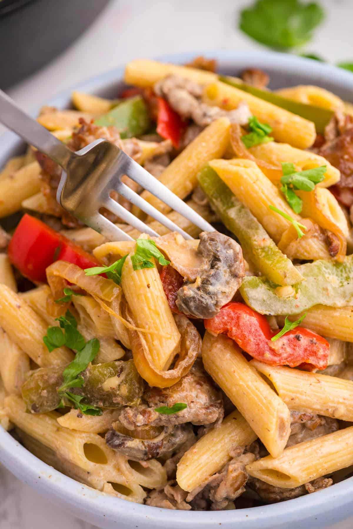 A close-up of a bowl of penne pasta with sautéed bell peppers, onions, mushrooms, and a creamy sauce, garnished with chopped parsley and a fork twirling some pasta.