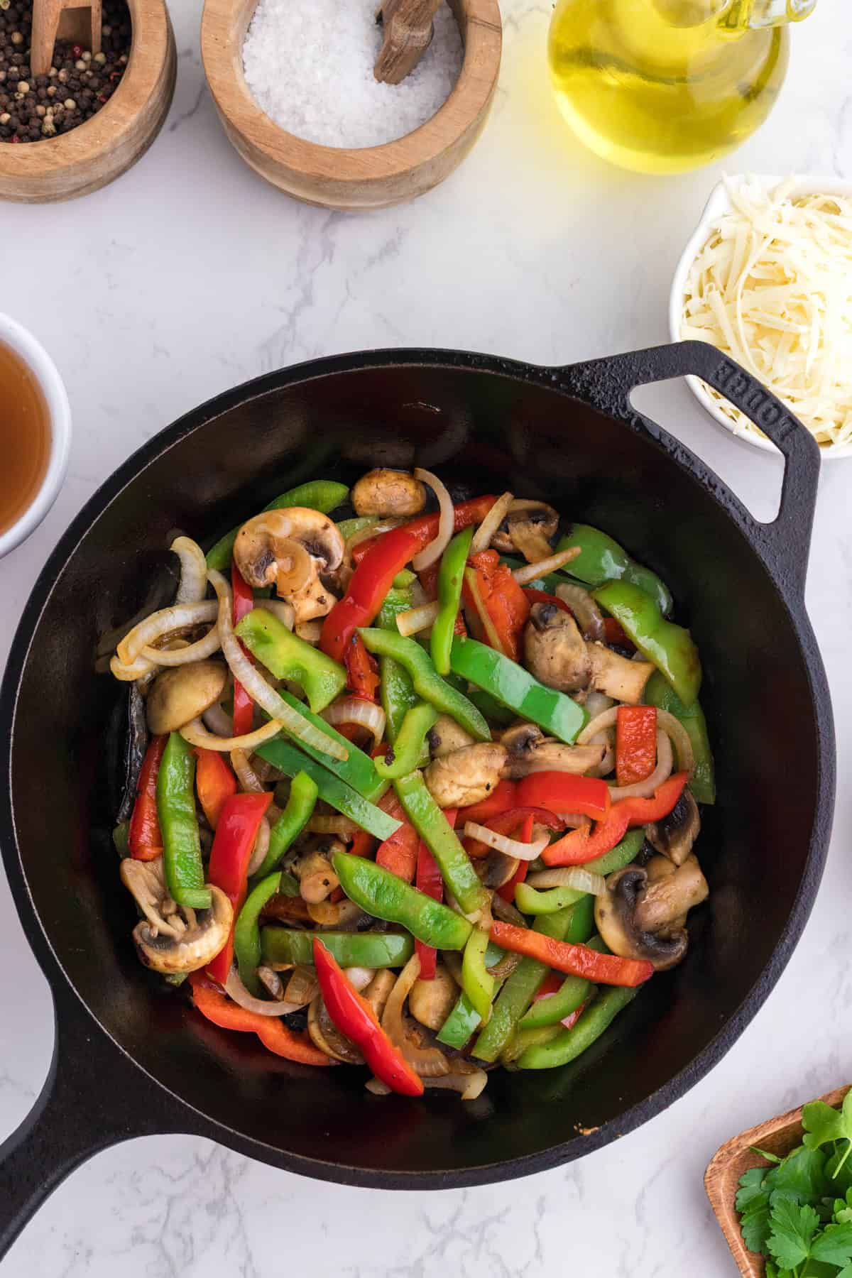 Sliced green and red bell peppers, onions, and mushrooms cooking in a black cast iron skillet on a white countertop surrounded by various ingredients.