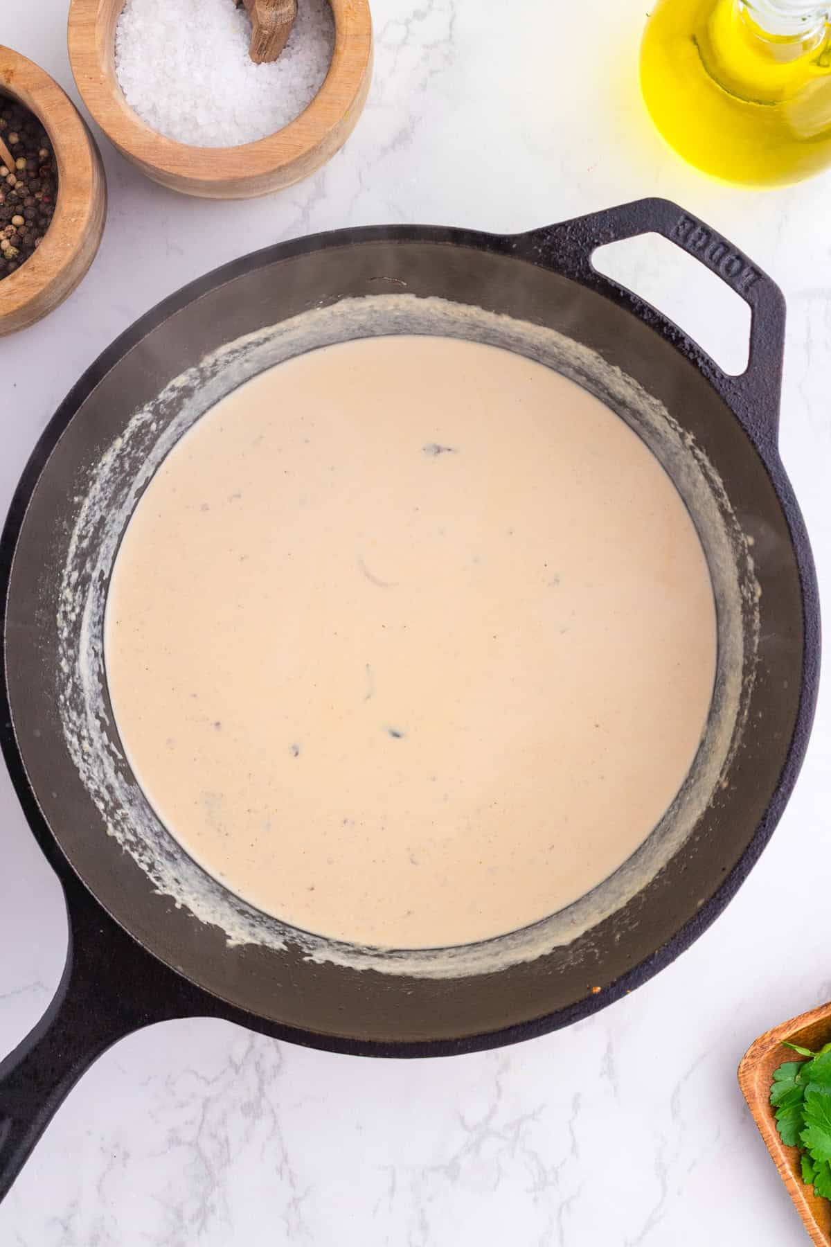 A cast iron skillet filled with a creamy beige sauce sits on a white countertop, surrounded by bowls of salt, pepper, and a bottle of olive oil.