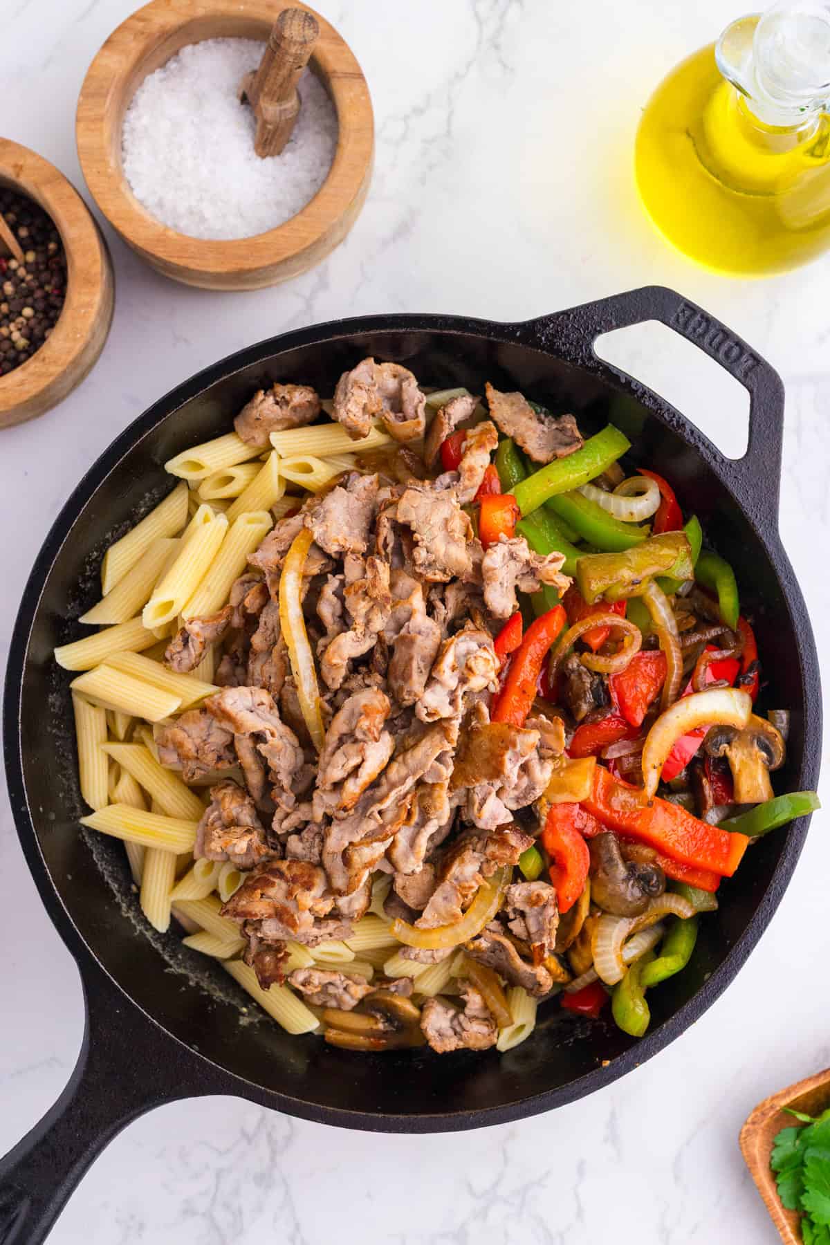 A skillet containing cooked penne pasta, sautéed sliced pork, and mixed vegetables including bell peppers, onions, and mushrooms. Salt, pepper, and olive oil are nearby on the counter.