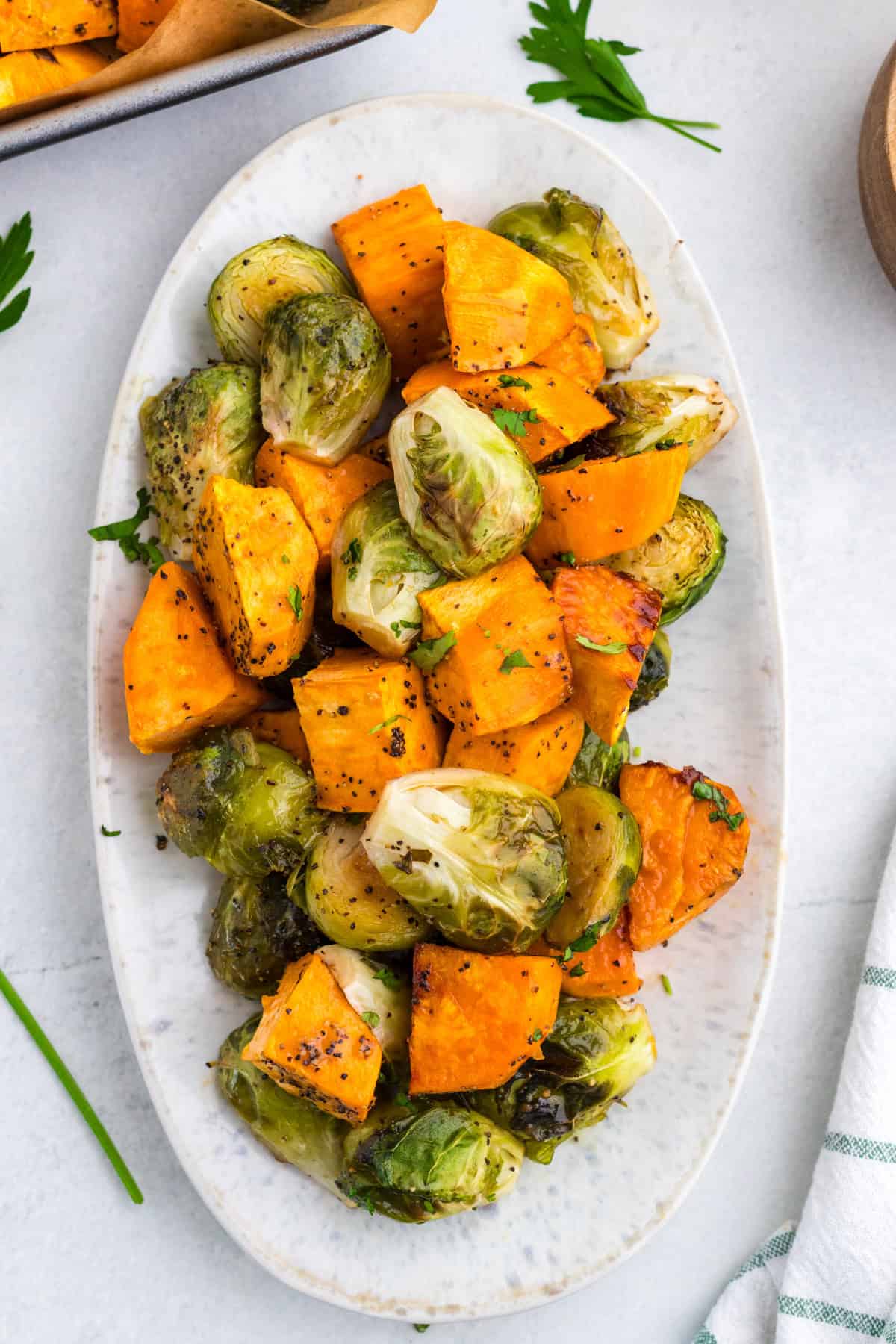 Roasted Brussels sprouts and sweet potato cubes seasoned with black pepper, served on an oval white plate, garnished with chopped parsley.