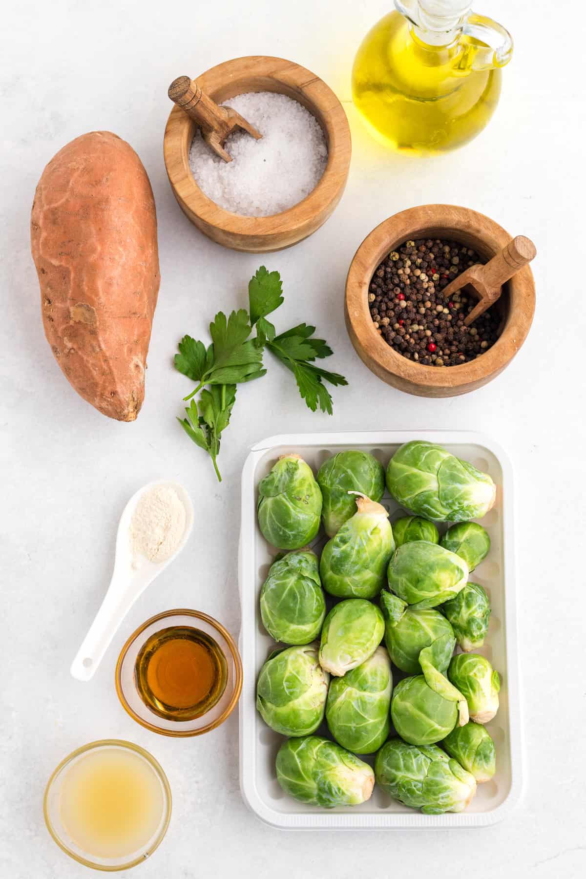 Tray of Brussels sprouts, a sweet potato, parsley, olive oil, salt, pepper, maple syrup, lemon juice, and garlic powder are arranged on a white surface.