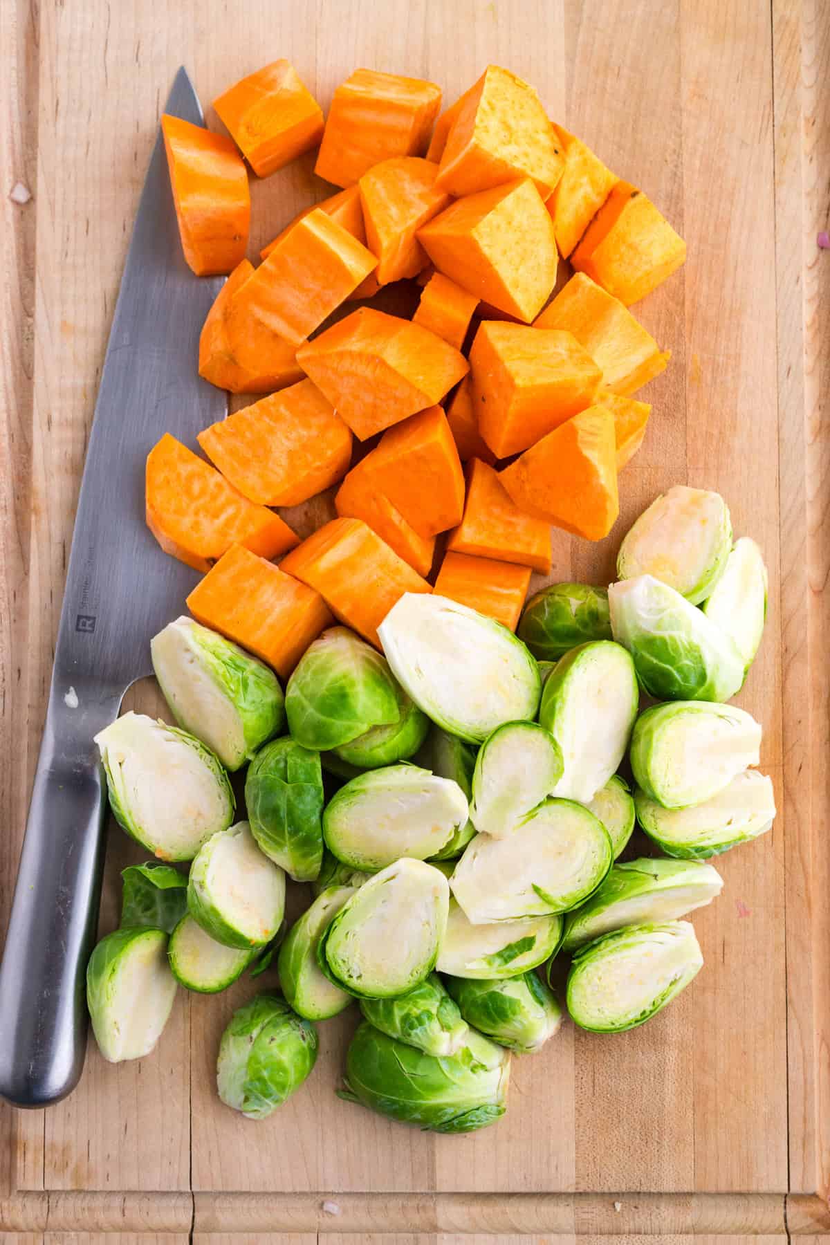 Chunks of sweet potato and halved Brussels sprouts are arranged on a wooden cutting board next to a large kitchen knife.
