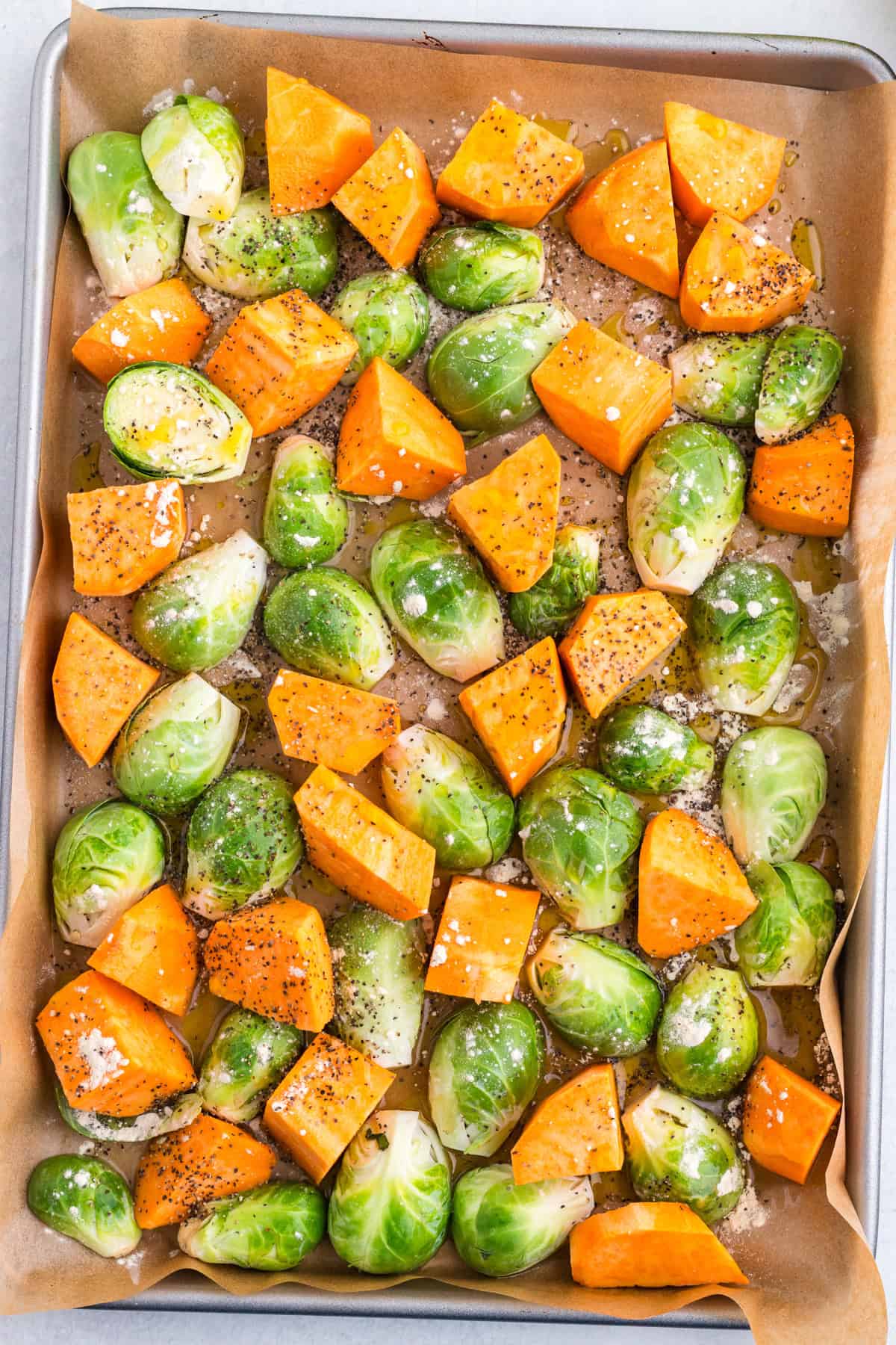 Chopped Brussels sprouts and sweet potatoes on a parchment-lined baking sheet, sprinkled with salt, pepper, garlic powder and olive oil.