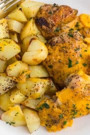 A plate with roasted potato cubes and baked chicken topped with melted cheddar cheese, garnished with chopped herbs, with a fork on the side.