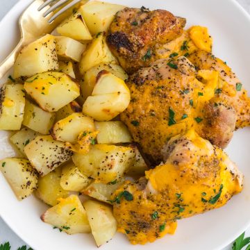 A plate with roasted potato cubes and baked chicken topped with melted cheddar cheese, garnished with chopped herbs, with a fork on the side.