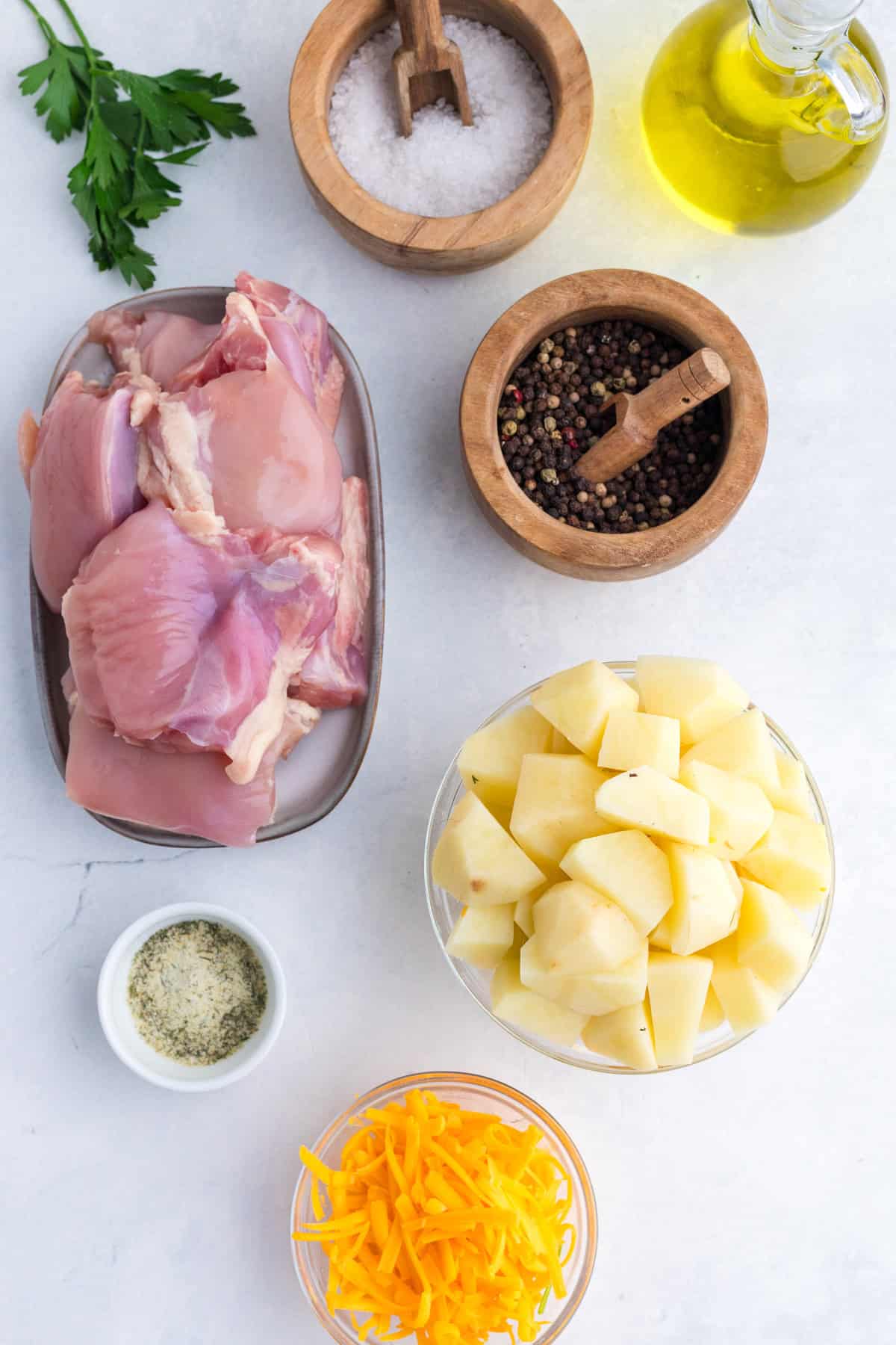 Raw chicken, peeled potatoes, shredded cheese, salt, pepper, olive oil, seasoning, and parsley are arranged on a white surface.