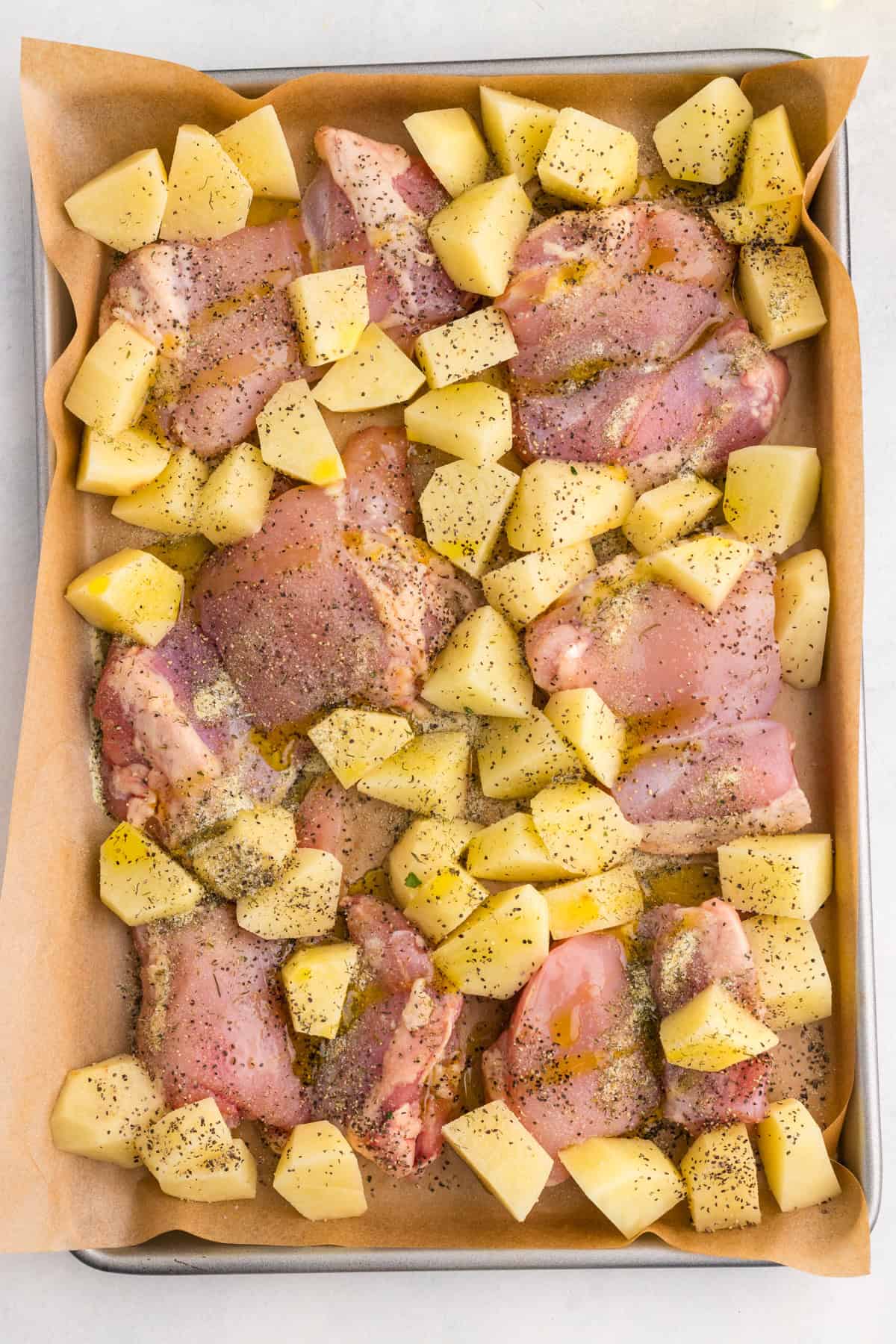 Raw chicken pieces and diced potatoes are spread on a parchment-lined baking sheet, seasoned with black pepper and herbs.