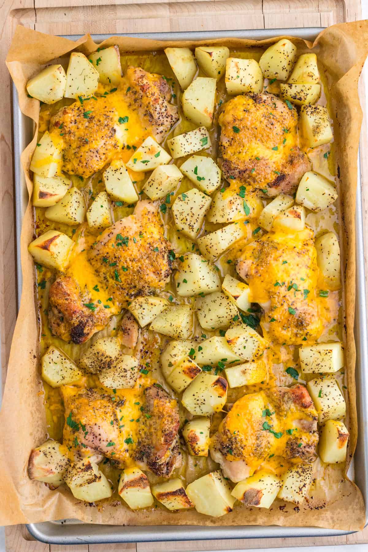 Sheet pan with baked chicken thighs topped with melted cheese, surrounded by seasoned roasted potato chunks, garnished with chopped parsley on parchment paper.