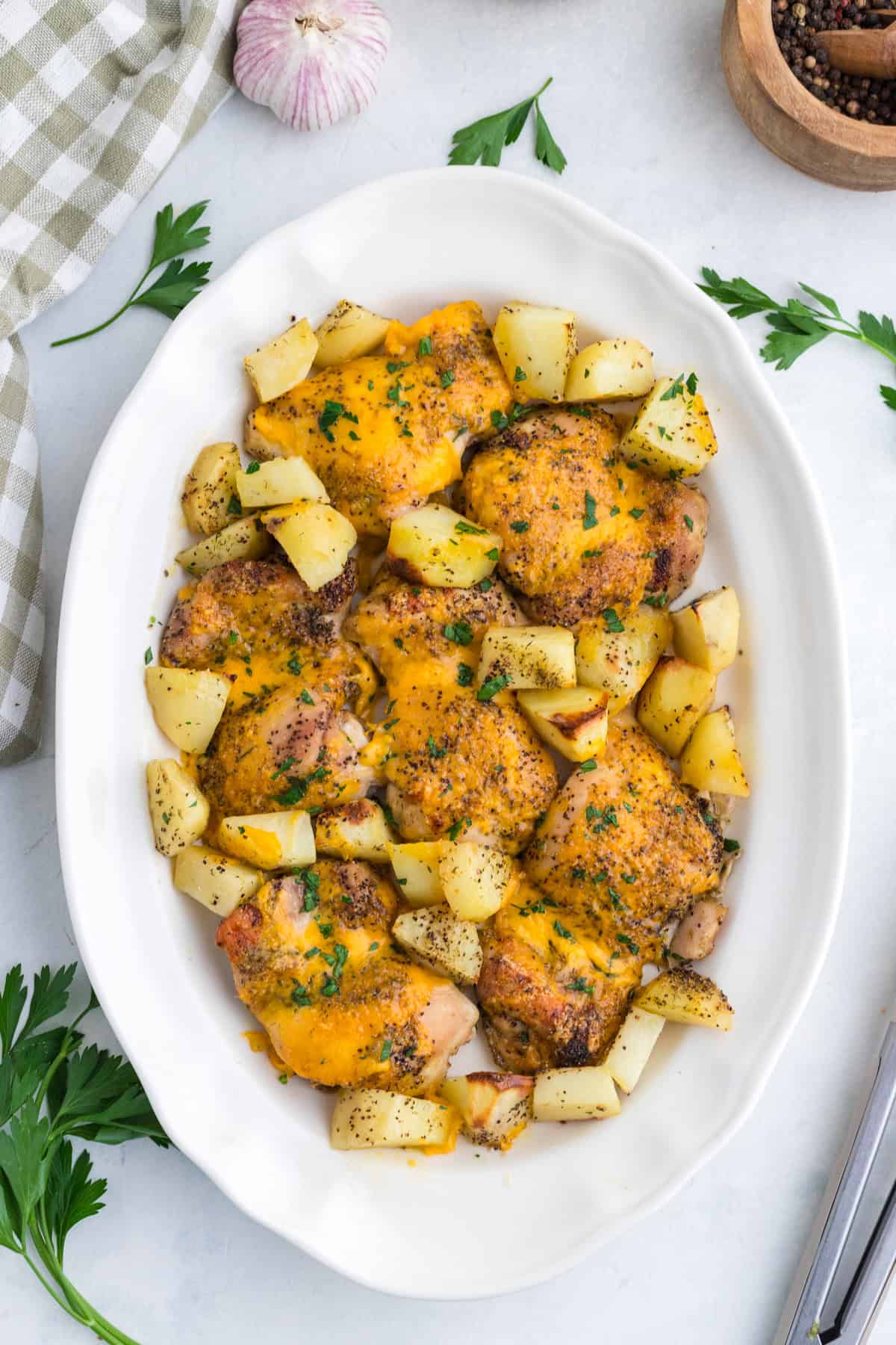 Baked chicken thighs topped with melted cheese, surrounded by roasted potato cubes, served on a white oval dish with fresh herbs sprinkled on top.