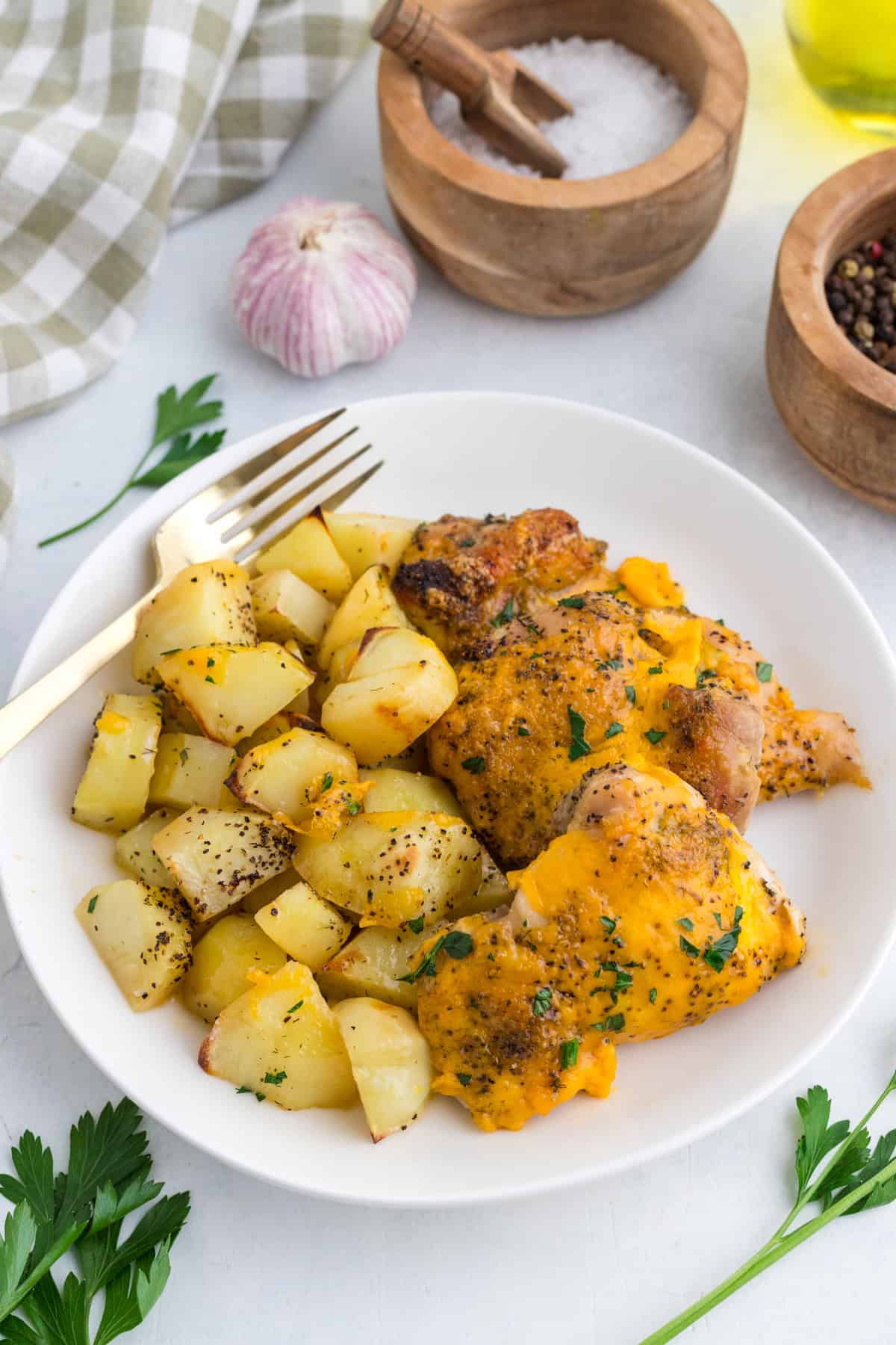 White plate with baked chicken topped with melted cheese and a side of roasted potatoes, garnished with parsley; a fork rests on the plate. Salt, pepper, and garlic are visible in the background.