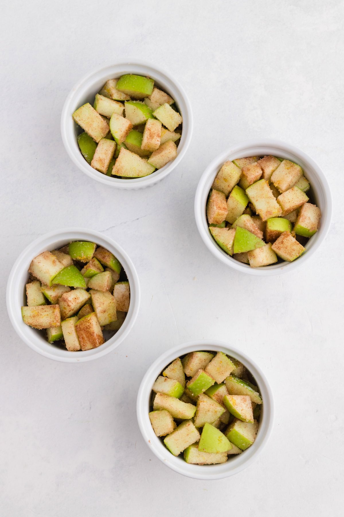 Quatre petits bols blancs remplis de dés de pommes vertes saupoudrés de cannelle, disposés sur un support clair.