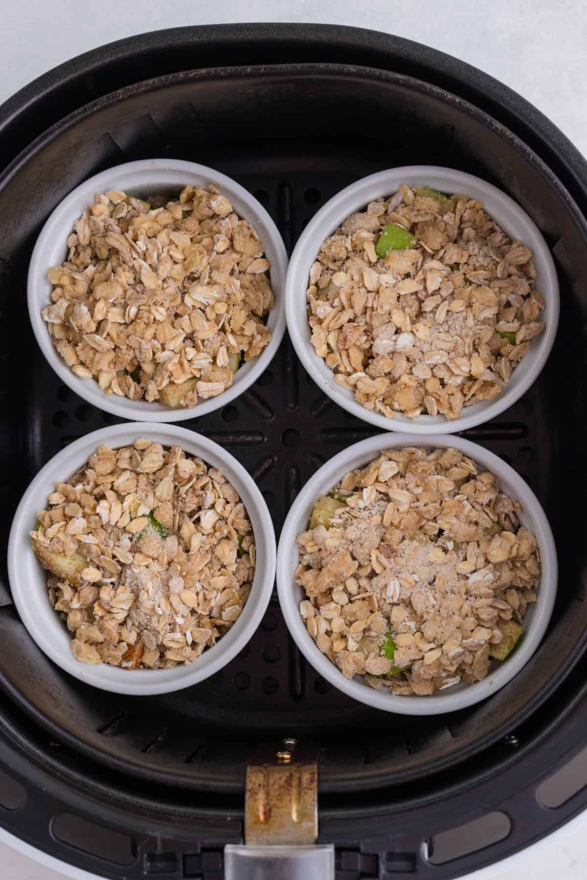 Quatre ramequins blancs remplis d'un crumble garni d'avoine sont disposés dans le panier d'une friteuse à air chaud, prêts à être cuits.
