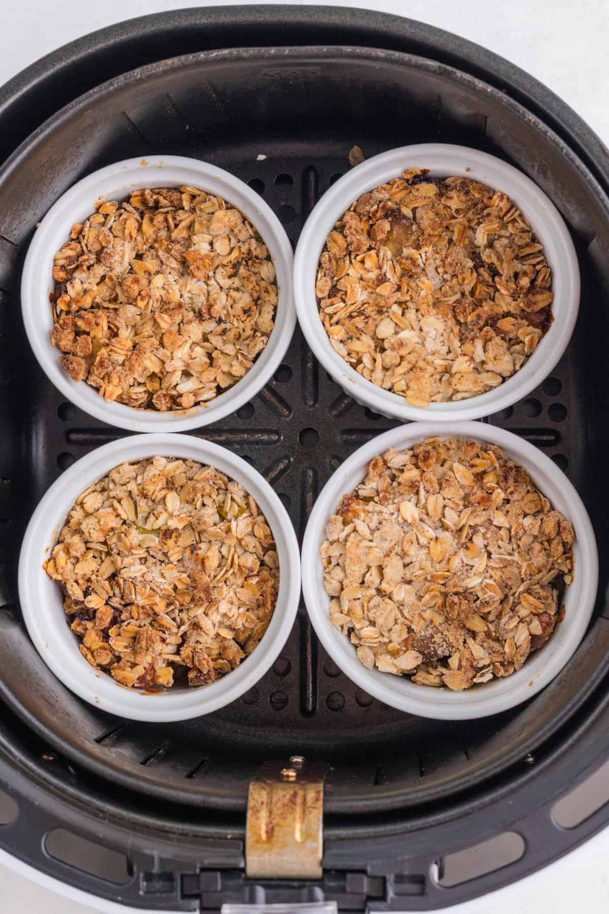 Quatre ramequins blancs remplis de crumble à l'avoine cuit au four sont disposés dans le panier d'une friteuse à air chaud.