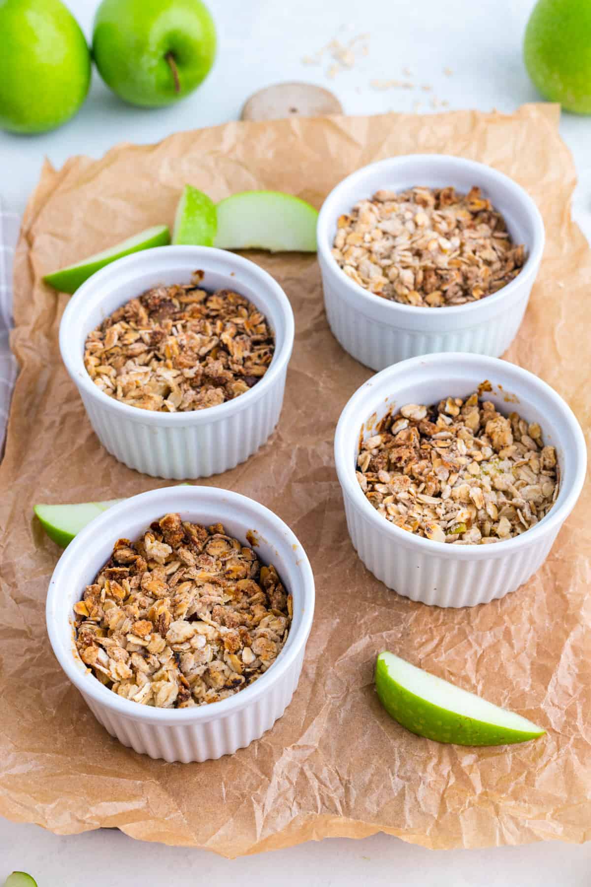 Quatre ramequins blancs remplis de crumble à l'avoine cuit au four sont posés sur du papier sulfurisé marron, entourés de tranches de pomme verte.