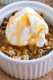 A white ramekin filled with oat crumble, topped with a scoop of vanilla ice cream and drizzled with caramel sauce.
