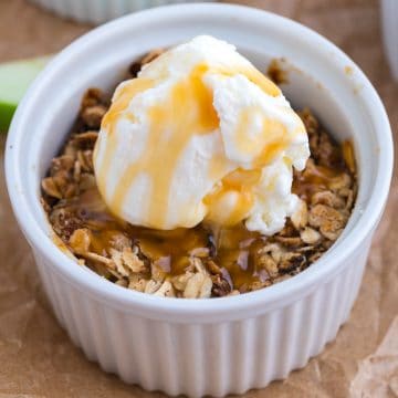 A white ramekin filled with oat crumble, topped with a scoop of vanilla ice cream and drizzled with caramel sauce.