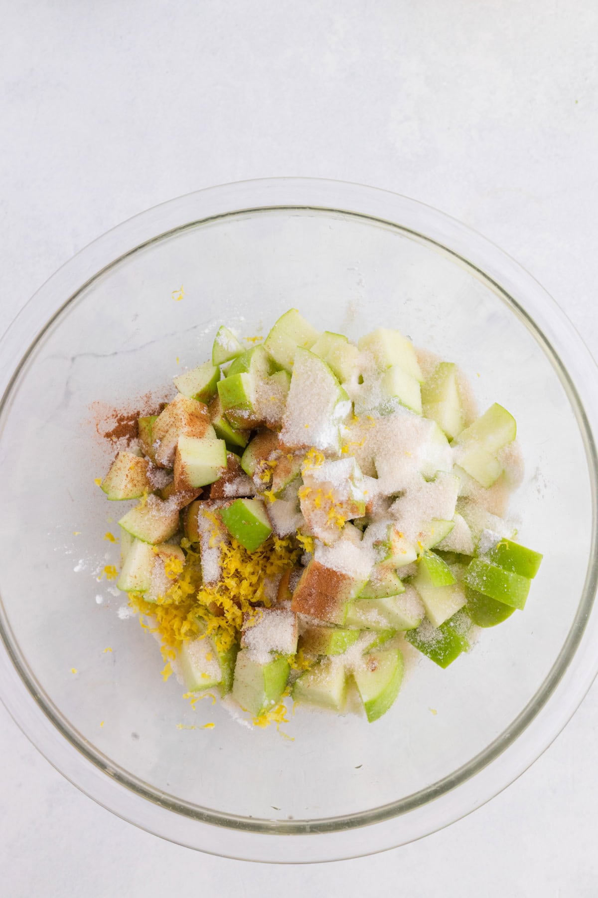 Des morceaux de pommes vertes dans un saladier en verre, saupoudrés de sucre, de cannelle et de zeste de citron.