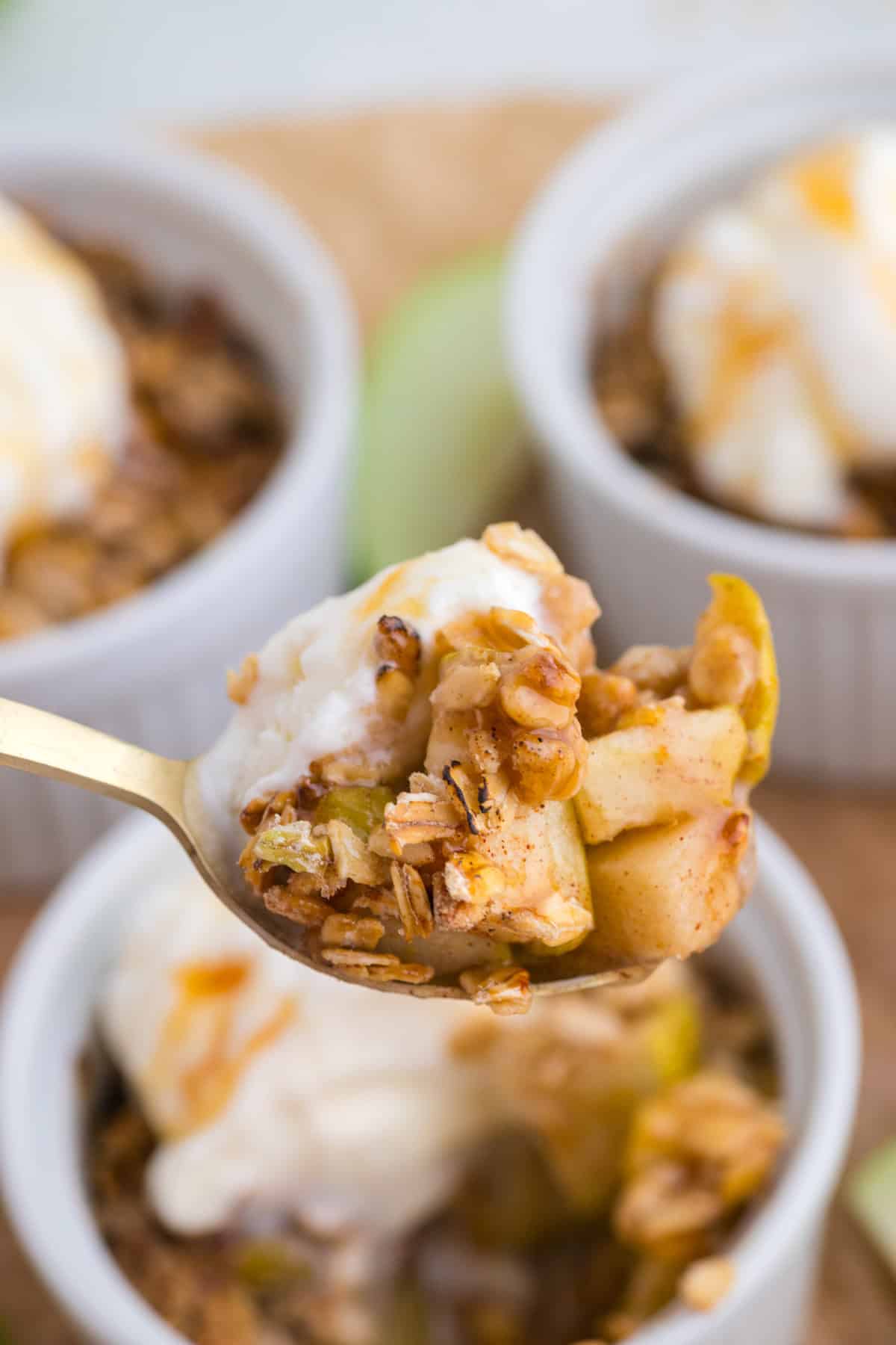 Une cuillerée de crumble aux pommes et aux flocons d'avoine, accompagnée d'une boule de glace à la vanille, posée au-dessus d'un ramequin, avec d'autres desserts en arrière-plan.