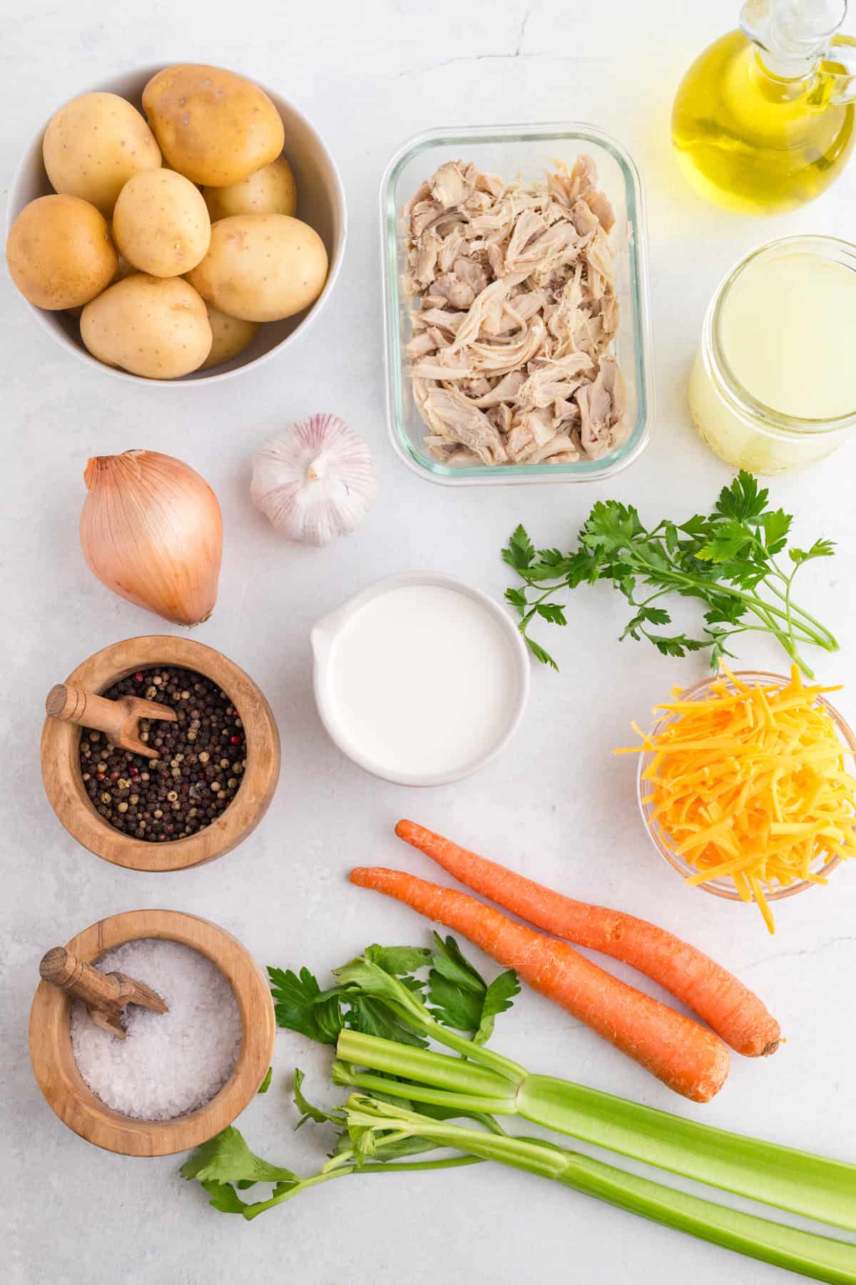 Top-down view of raw potatoes, shredded chicken, oil, broth, garlic, onion, parsley, black pepper, salt, cream, shredded cheese, carrots, and celery arranged on a light surface.