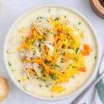 A bowl of creamy soup with shredded chicken, potatoes, carrots, chopped herbs, and shredded cheddar cheese on top, placed on a light surface.