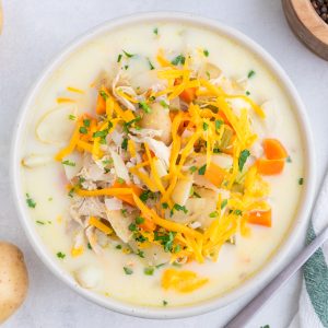 A bowl of creamy soup with shredded chicken, potatoes, carrots, chopped herbs, and shredded cheddar cheese on top, placed on a light surface.