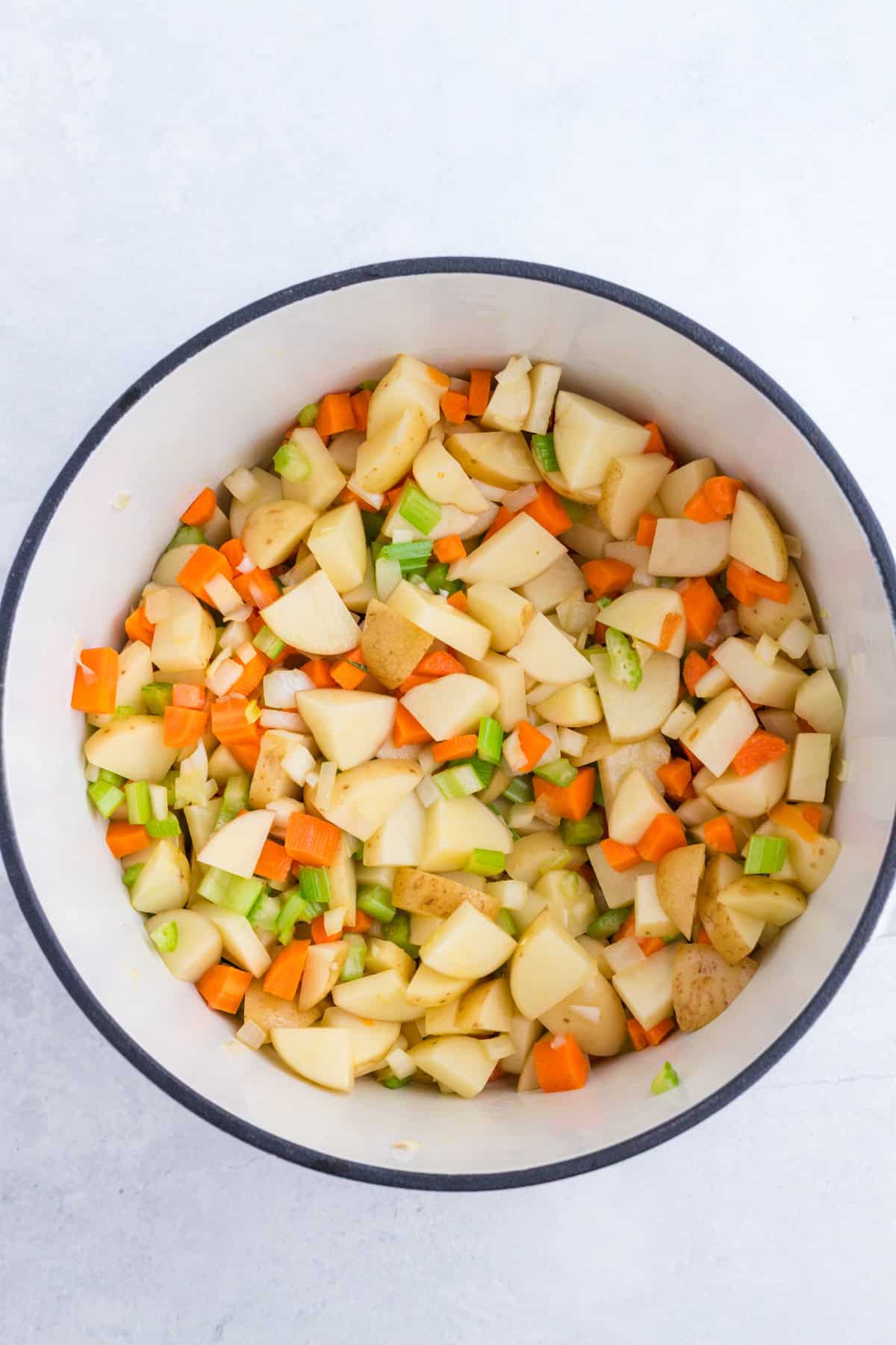 A white pot filled with chopped potatoes, carrots, celery, and onions on a light surface.
