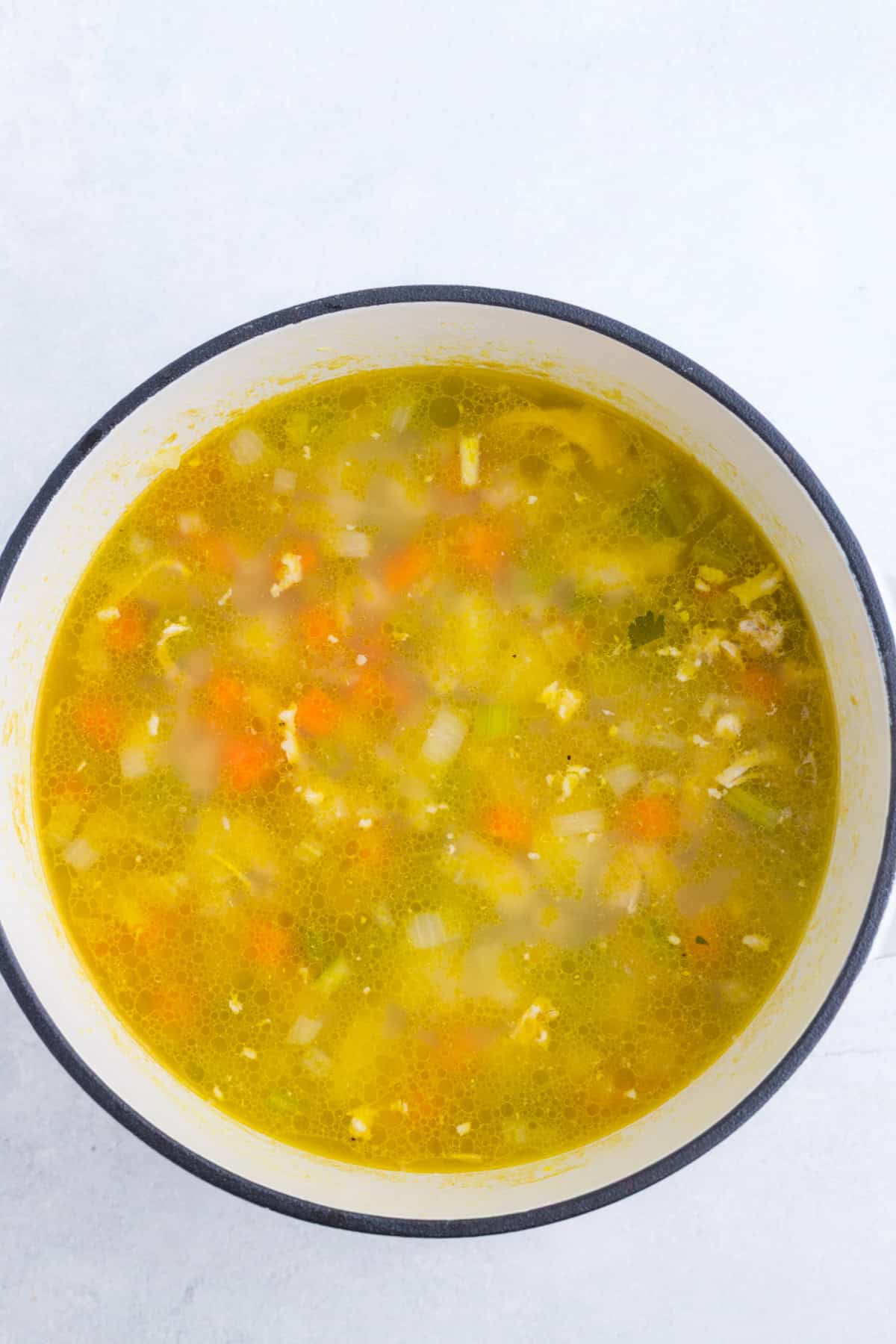 A large pot filled with yellow broth containing chopped vegetables such as carrots, celery, and onions, seen from above against a light background.