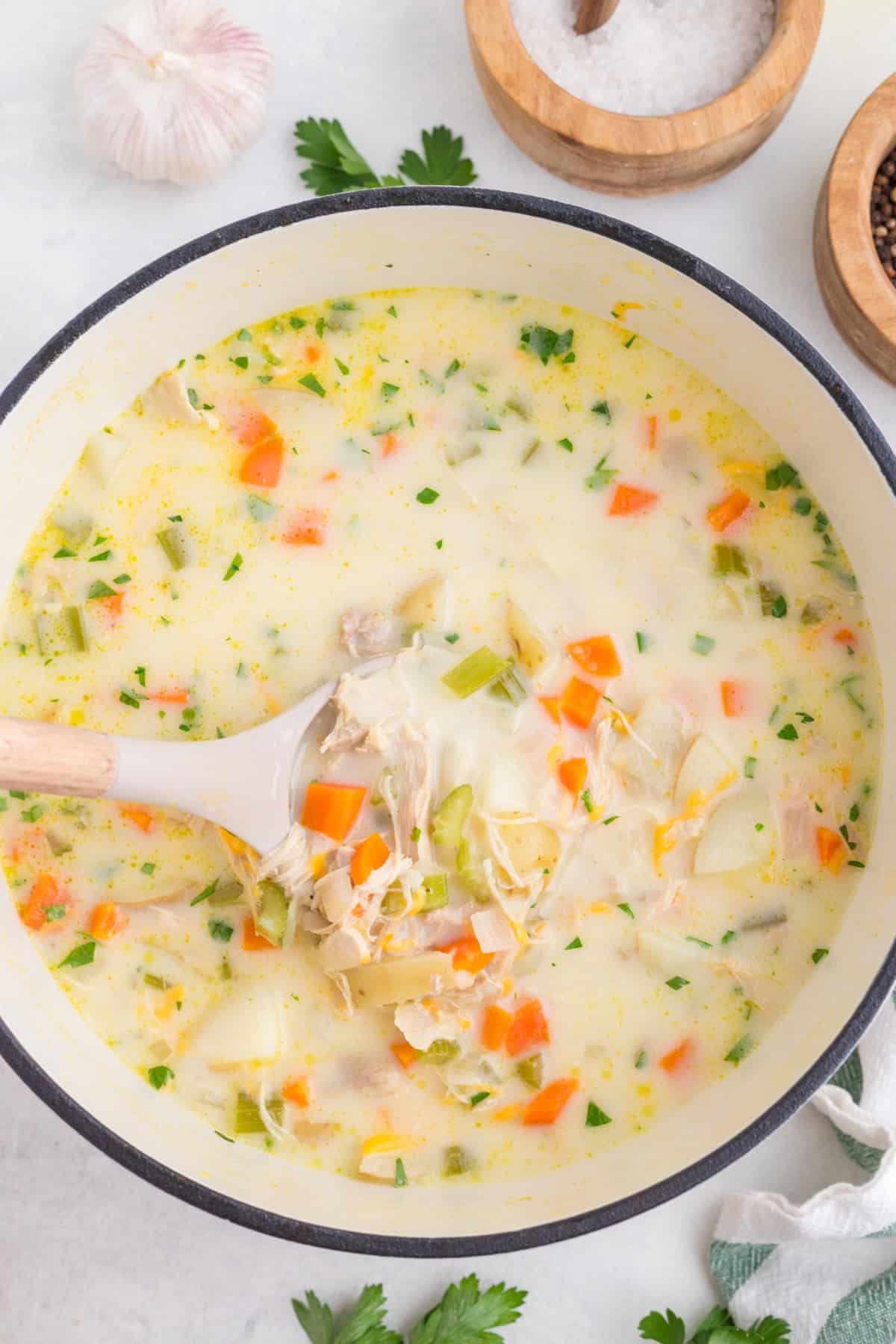 A pot of creamy chicken soup with chunks of chicken, carrots, celery, potatoes, and herbs, being stirred with a white ladle.