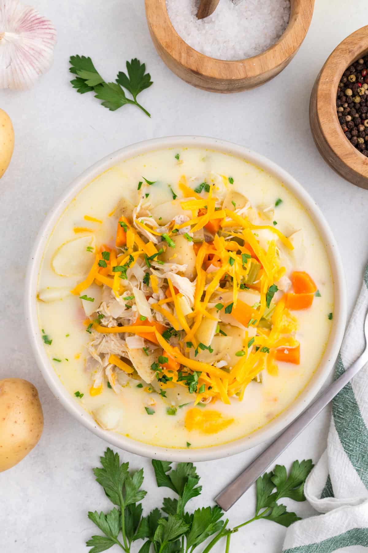 Bowl of creamy chicken potato soup topped with shredded cheese and herbs, surrounded by parsley, potatoes, salt, pepper, and a garlic bulb on a white surface.