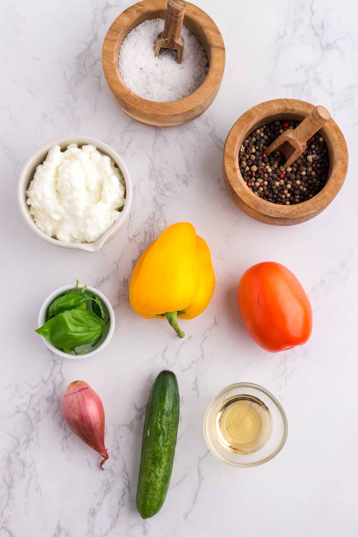 Assorted ingredients on a marble surface: salt, pepper, cottage cheese, yellow bell pepper, tomato, fresh basil, shallot, cucumber, and a small glass of oil.