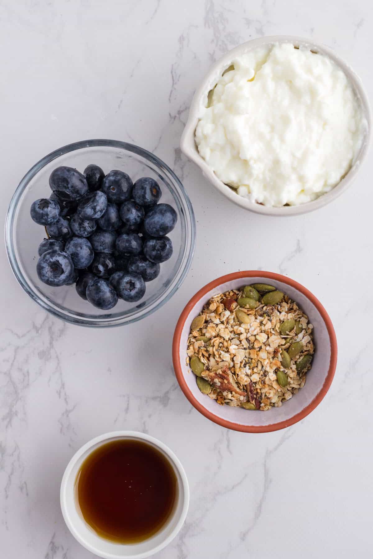 Four bowls containing blueberries, cottage cheese, granola, and a small amount of dark liquid, likely syrup, are arranged on a white marble surface.