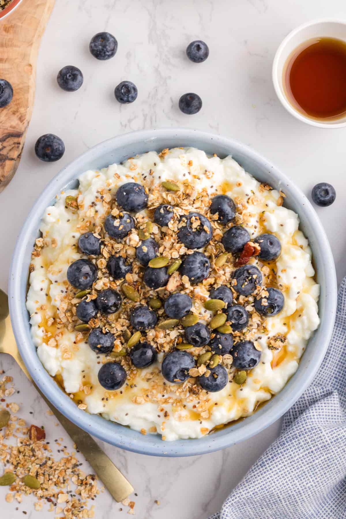 A bowl of cottage cheese topped with fresh blueberries, granola, and seeds sits on a white surface, with a small cup of syrup and scattered blueberries nearby.