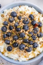 A bowl of cottage cheese topped with blueberries, granola, seeds, and a drizzle of honey sits on a light surface.