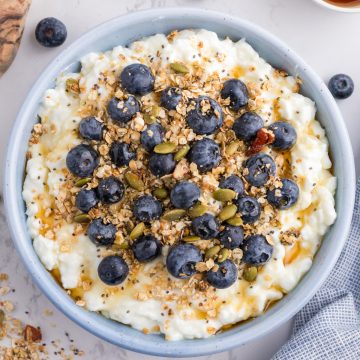 A bowl of cottage cheese topped with blueberries, granola, seeds, and a drizzle of honey sits on a light surface.