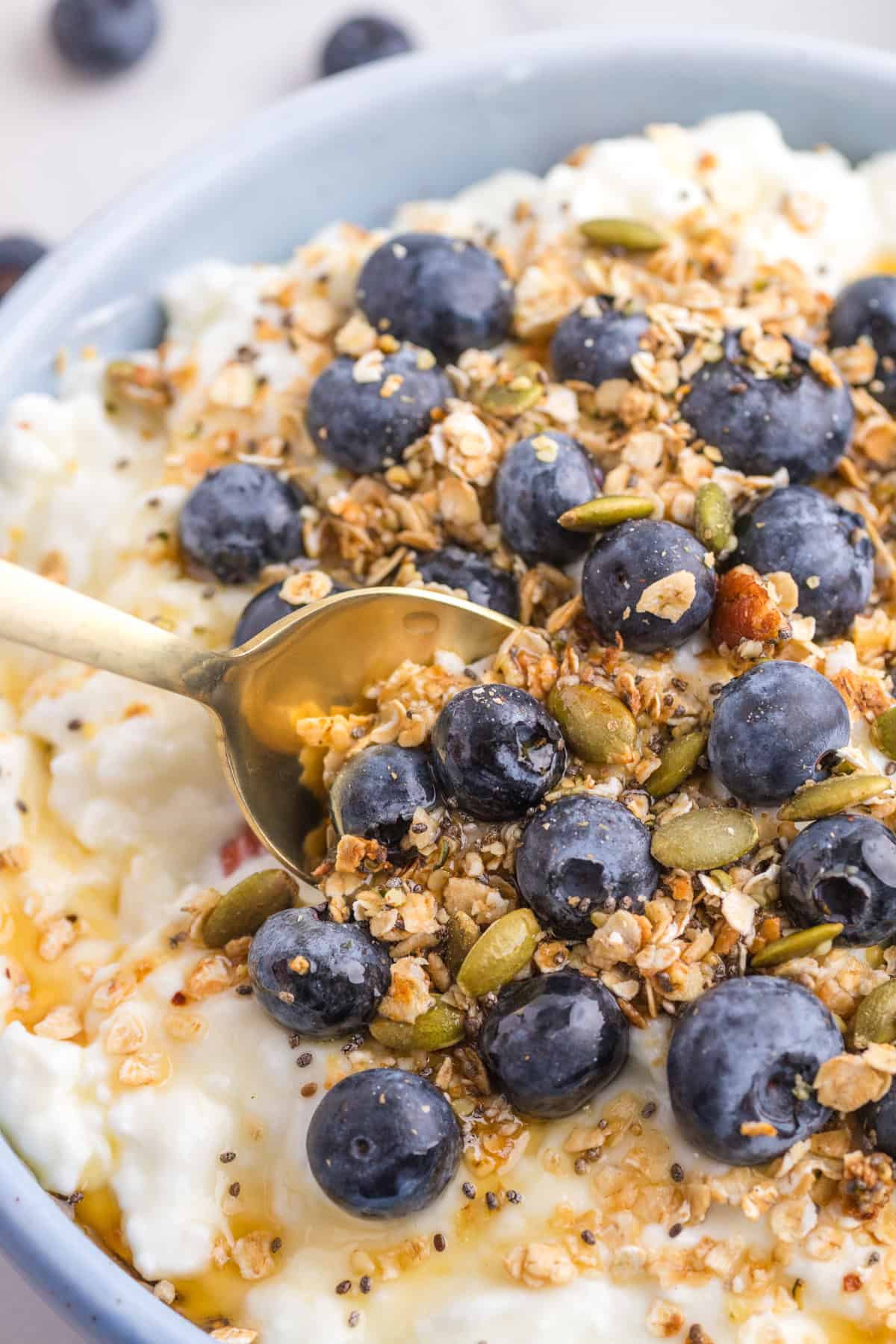 Close-up of a bowl with cottage cheese, blueberries, granola, pumpkin seeds, and a drizzle of maple syrup. A gold spoon is scooping the mixture.
