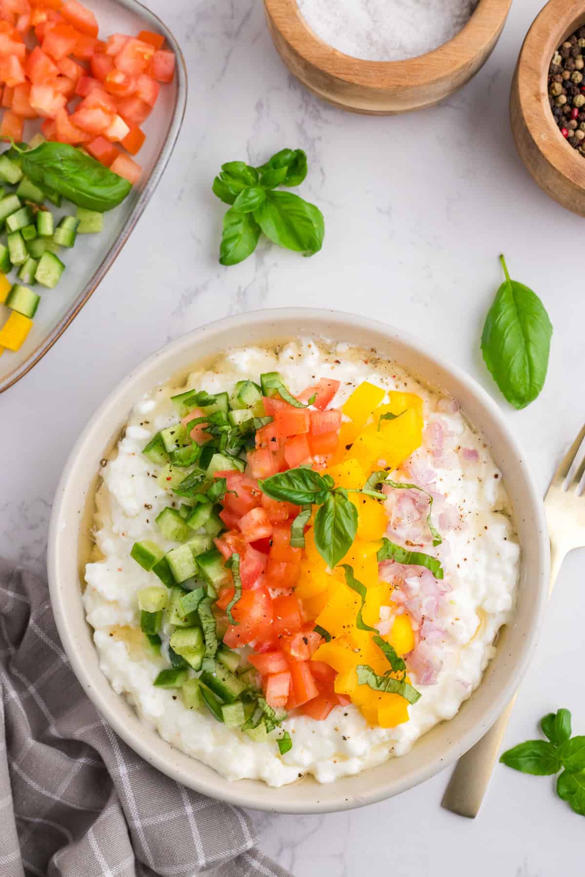 A bowl of cottage cheese topped with diced tomatoes, cucumbers, yellow peppers, and onions, garnished with basil, next to bowls of salt, pepper, and extra vegetables.