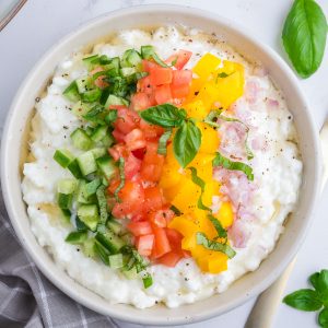 A bowl of cottage cheese topped with chopped cucumber, tomato, yellow bell pepper, red onion, basil, and black pepper.