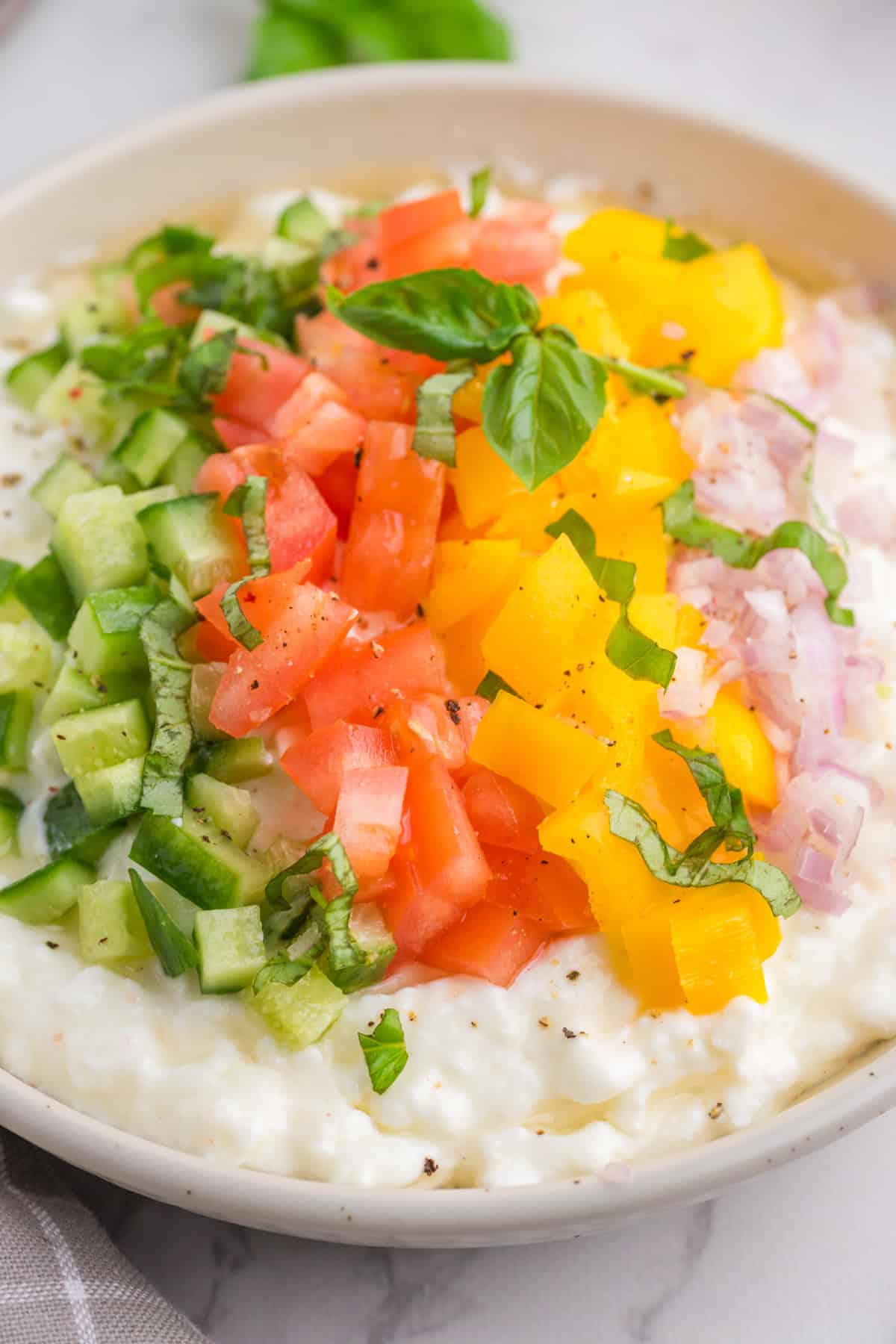 A bowl of cottage cheese topped with diced cucumber, tomato, yellow bell pepper, shallot, and fresh basil, with cracked black pepper.