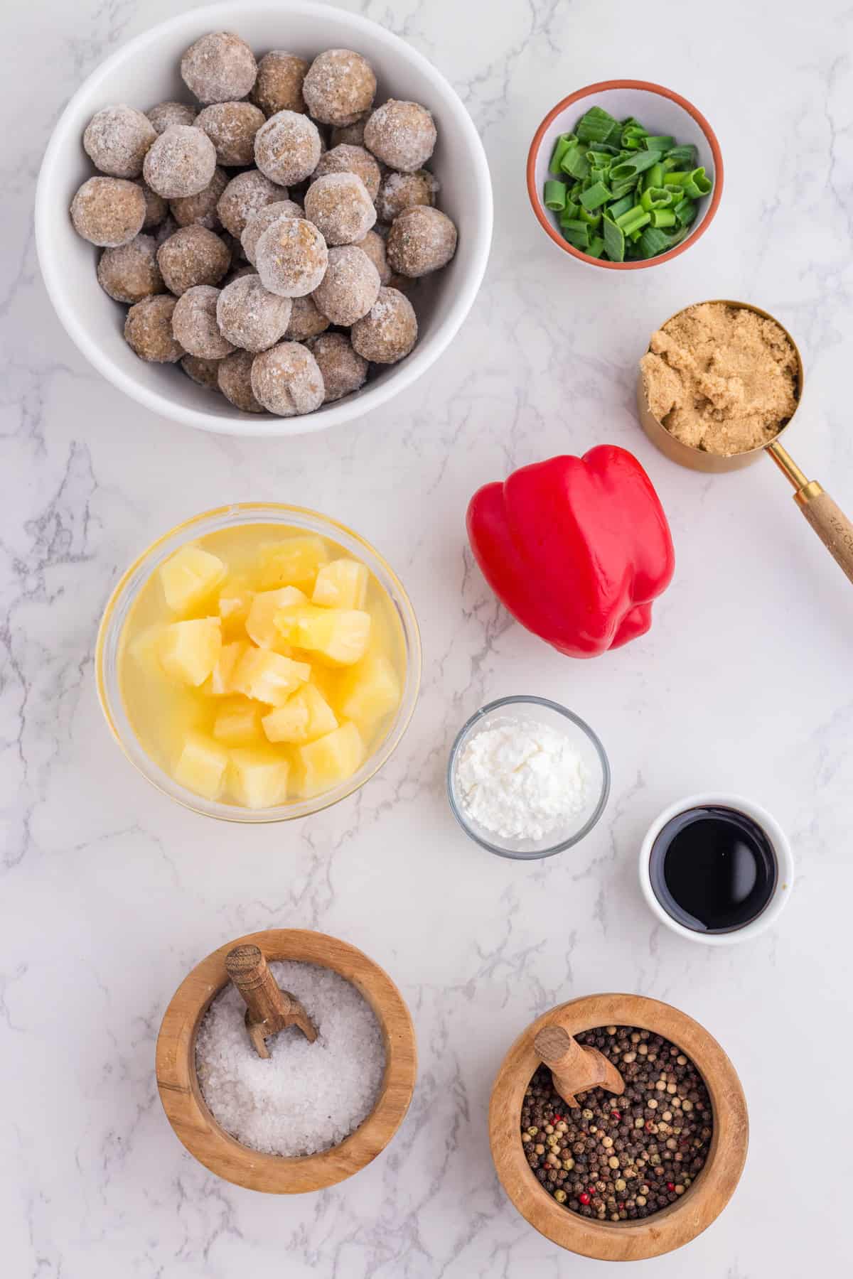 Overhead view of bowls containing frozen meatballs, chopped green onions, brown sugar, pineapple chunks, sliced red bell pepper, cornstarch, soy sauce, salt, and pepper on a white surface.