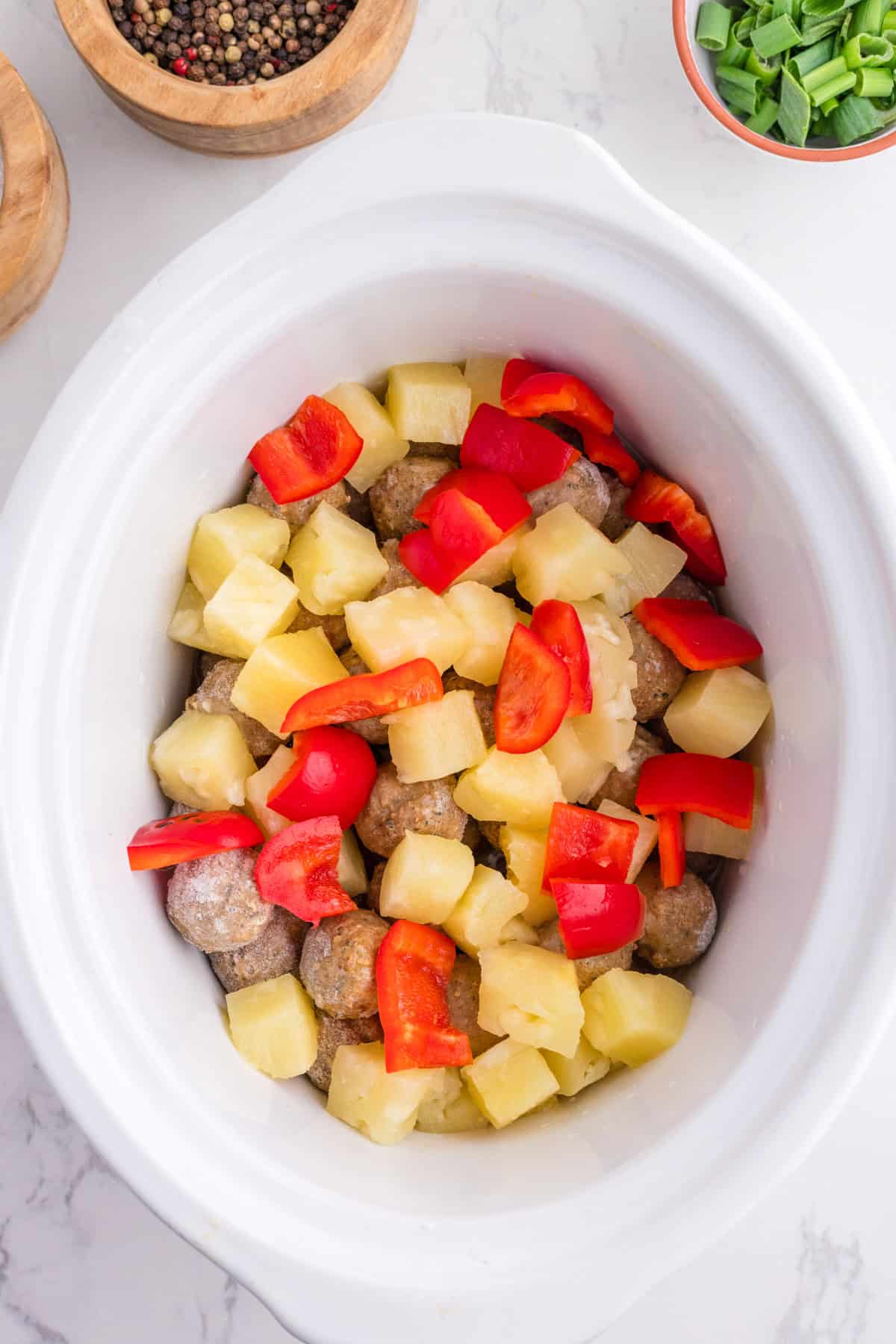Crockpot filled with meatballs, pineapple chunks, and red bell pepper pieces, viewed from above on a white surface with seasonings nearby.