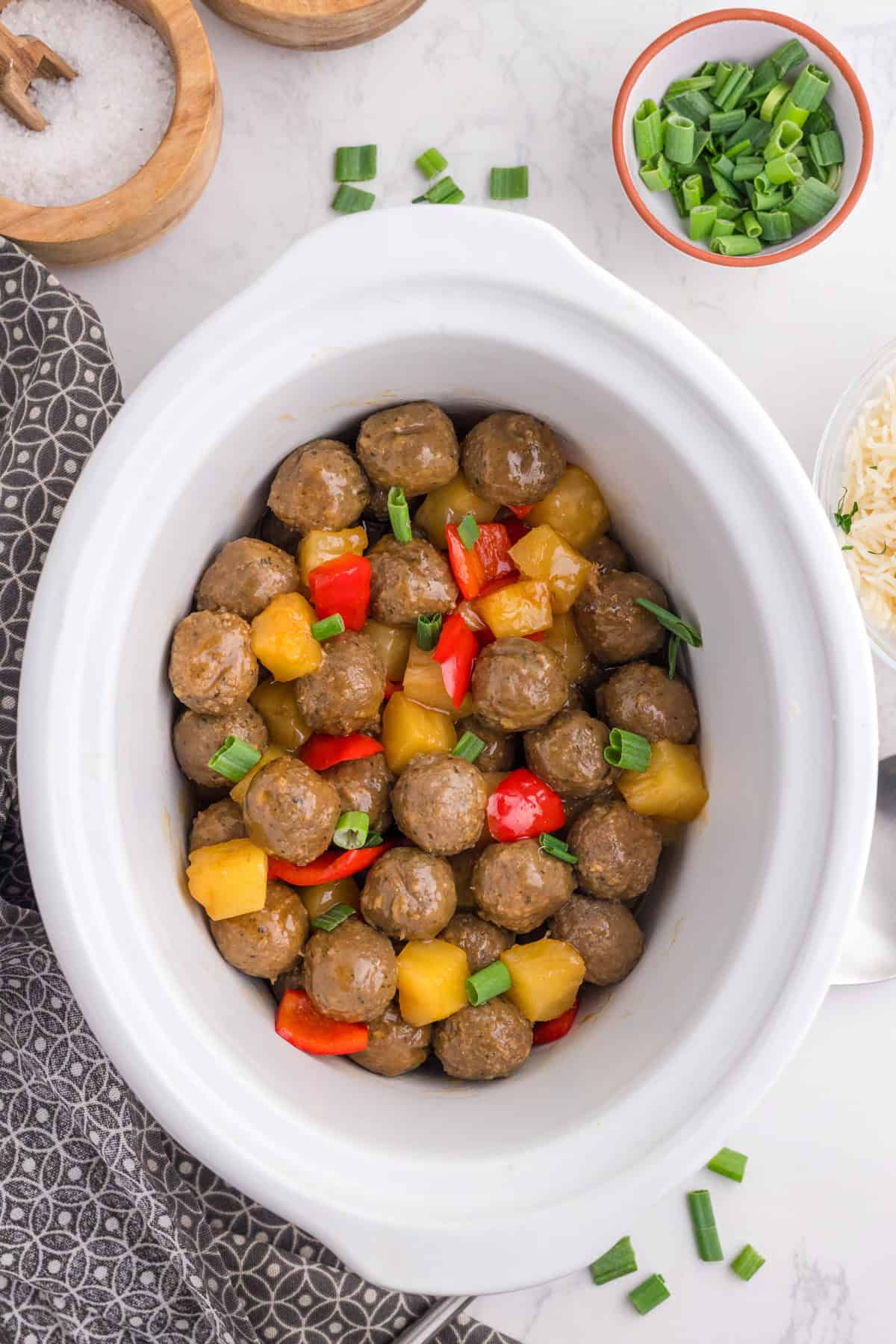 Meatballs, pineapple chunks, and red bell peppers in a white slow cooker, garnished with chopped green onions.