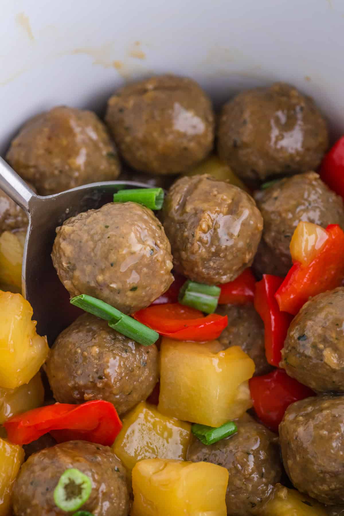 Meatballs with pineapple chunks, red bell peppers, and sliced green onions, served in a bowl with a serving spoon.