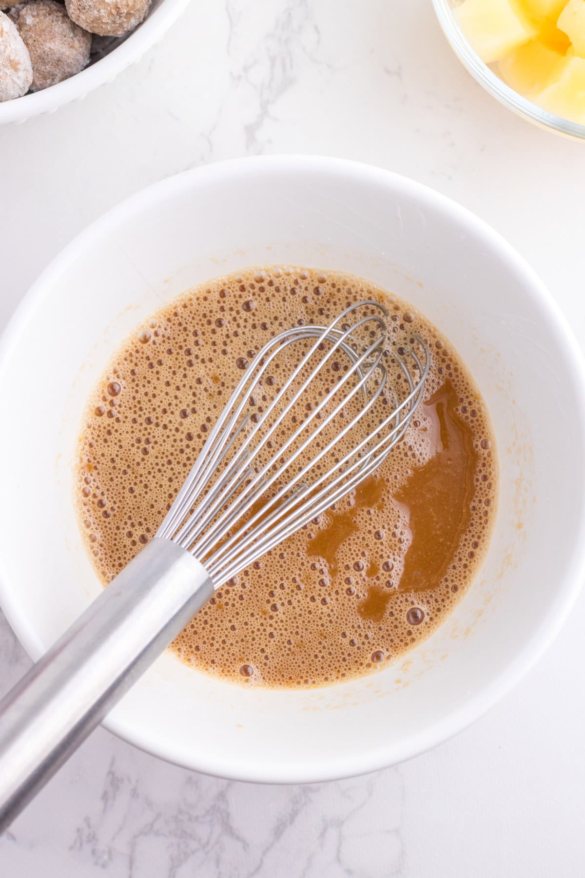 A metal whisk rests in a white bowl containing a frothy brown liquid mixture, with ingredients visible nearby on a white marble surface.