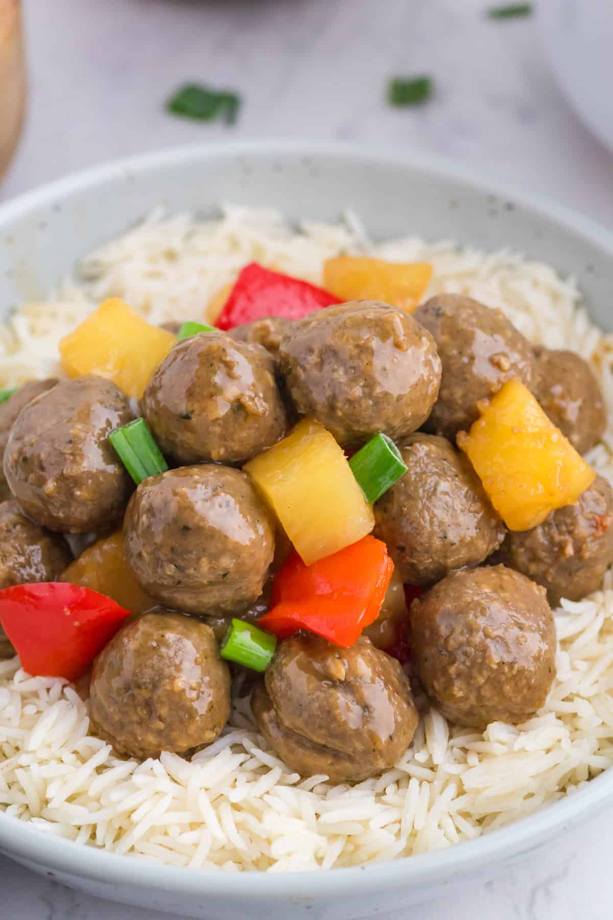Meatballs with pineapple chunks, red bell pepper, and green onions served over a bed of white rice in a light gray bowl.
